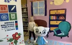 Na tle bajkowego pokoju przy Kici Koci stoi uśmiechnięta dziewczynka w blond włosach. Z lewej strony stoi logo biblioteki.