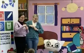 Z przodu przy Kici Koci stoją dwie uśmiechnięte Panie trzymając coś małego w dłoniach. Z lewej strony stoi logo biblioteki.