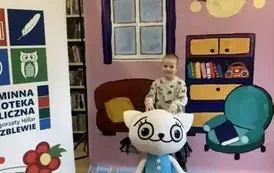 Na tle bajkowego pokoju przy Kici Koci stoi chłopiec w jasnej bluzie uśmiechając się.