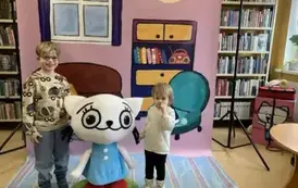 Na tle bajkowego pokoju przy Kici Koci stoi dziewczynka i chłopiec.