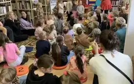 Dzieci, opiekunowie i rodzice siedzą na dywanie i patrzą na Bajankę, kt&oacute;ra na tle bajkowego pokoju trzyma w ręku rekwizyty. Po lewej stronie stoi logo biblioteki.