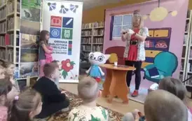 Na tle bajkowego pokoju obok Kici Koci stoi przy stoliku Bajanka w okularach i rękawiczkach robiąc eksperyment. Po lewej stronie stoi dziewczynka i patrzy. 