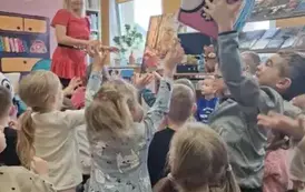 Dzieci na dywanie podają do siebie z wyciągniętymi rączkami do g&oacute;ry duży mięki klocek Kici Koci. Z przodu stoi Bajanka i patrzy.