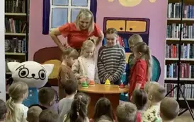 Na tle bajkowego pokoju przy stoliku obok Kici Koci stoi czw&oacute;rka dzieci, kt&oacute;ra patrzy na rekizyty położone na stoliku. Nad nimi pochylona jest Bajanka bohaterka bajki.