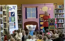 Dzieci siedzą na siedziskach, na wprost wisi tło bajkowego pokoju w kt&oacute;rym stoi Bajanka uczestniczka bajki z Kicią Kocią. Po prawej stronie stoją duże klocki Kici Koci.