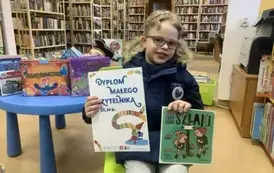 Chłopiec w blond włosach i okularkach siedzi na krzesłku trzymając dyplom i wypożyczoną książeczkę.