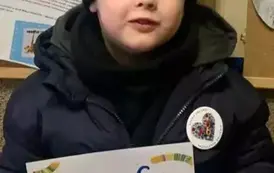 Chlopiec w czarnej czapce stoi przy korkowej tablicy z grafikami dotyczącymi akcji MAŁA KSIĄŻKA WIELKI CZŁOWIEK. Trzyma w rękach dyplom.