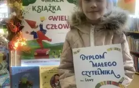 Dziewczynka w blond włosach stoi przy tablicy z plakatem dotyczącym akcji MAŁA KSIĄŻKA WIELKI CZŁOWIEK. Trzyma w rękach dyplom.