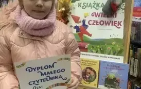Dziewczynka stoi przy tablicy z plakatem dotyczącym akcji MAŁA KSIĄŻKA WIELKI CZŁOWIEK. Trzyma w rękach dyplom i kartę Małego Czytelnika.