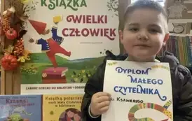Chłopiec w ciemnych włosach stoi przy tablicy z plakatem MAŁA KSIĄŻKA WIELKI CZŁOWIEK. Trzyma dyplom.
