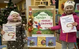 Dwie uśmiechnięte dziewczynki stoją przy korkowej tablicy z plakatem dotyczącym akcji. Jedna z dziewczynek wskazuje długopisem napis na plakacie MAŁA KSIĄŻKA WIELKI CZŁOWIEK. Z lewej strony stoi choinka.