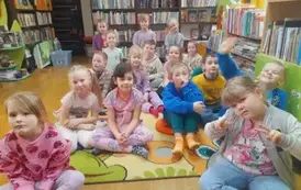 Dzieci siedzą na dywanie i słuchają bajki, kt&oacute;rą czyta im bibliotekarka.