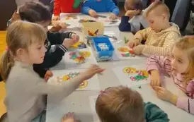 Dzieci przy białym stole obklejają plasteliną na kartkach kolorowe koguciki.