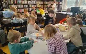 Dzieci przy stołach obklejają na kartkach plasteliną kolorowe koguciki. Przy jednym ze stoł&oacute;w stoi Pani z Przedszkola.