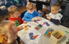 Chłopcy przy białym stole obklejają plasteliną na kartkach kolorowe koguciki.