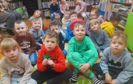 Dzieci siedzą na dywanie i słuchają bajki, kt&oacute;rą czyta im bibliotekarka.