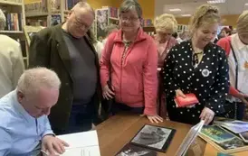 Przy biurku Janusz Szalewski podpisuje zakupione książki Czytelnikom, kt&oacute;rzy stoją w kolejce.