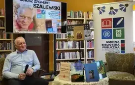 Autor siedzi na fotelu z mikrofonem w ręku, obok jest okrągły stolik z wystawionymi Jego książkami. Za autorem jest duży ekran z wyświetlonym plakatem dotyczącym spotkania,a po prawej stronie wisi logo biblioteki.