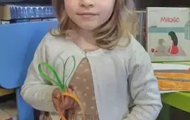 Dziewczynka w blond włosach do ramion siedzi na krzesełku i pokazuje swoją marchewkę z papieru.