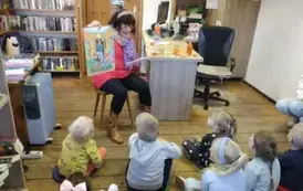 Dzieci siedzą na siedziskach, prowadząca siedzi na krześle czytając tekst z książki,a w prawej ręce pokazuje duży obrazek przedstawiający bawiących się dzieci na balu z balonami, na kt&oacute;rym odwiedził ich Pan Polcjant. 