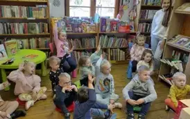 Dzieci siedzą na siedziskach, niekt&oacute;re z nich podnoszą rękę w celu zabrania głosu. Z prawej strony z tyłu stoi ich wychowawczyni.