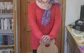 Pani prowadząca w czerwonym sweterku i okularach opiera się o drewniane krzesło i coś m&oacute;wi.