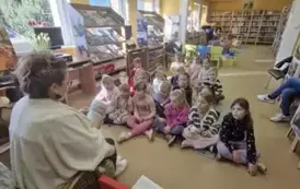 Dzieci siedzą na dywanie i siedziskach i patrzą na bibliotekarkę, kt&oacute;ra im czyta bajkę.