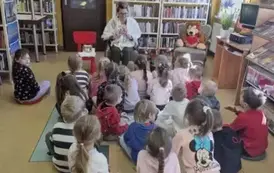 Dzieci siedzą na dywanie i siedziskach, z przodu na krzesełku siedzi bibliotekarka i uśmiechając się pokazuje dzieciom książeczkę pt "Calineczka"