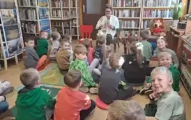 Dzieci siedzą na dywanie i siedziskach, z przodu na krzesełku siedzi bibliotekarka i uśmiechając się pokazuje dzieciom książeczkę pt "Calineczka"