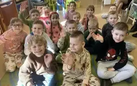 Dzieci siedzą na siedziskach w bibliotece, uśmiechają się, niekt&oacute;re podnoszą rączkę do g&oacute;ry