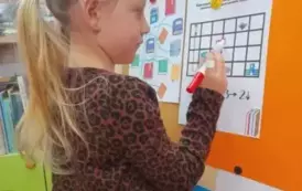 Dziewczynka w blond włosach i w kitkach stoi przy tablicy, na kt&oacute;rej doczepiona jest kartka z zadaniem. Dziewczynka rysuje pisakiem w kwadratach drogę chłopca do biblioteki.