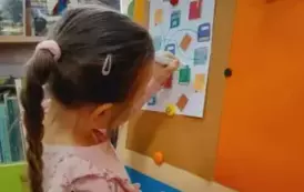 Dzieczynka w warkoczu stoi przy tablicy na kt&oacute;rej jest doczepiona kartka z zadaniem. Dziewczynka łączy pisakiem te same książki.
