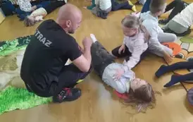 Dzieci leżą dobrane dw&oacute;jkami na podłodze i ćwiczą- przewracanie poszkodowanego na bok.