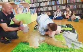 Dziewczyna robi sztuczne oddychanie fantomowi, obok siedzi prezes i się uśmiecha.