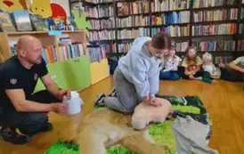 Dziewczyna w blond włosach i okularach uciska klatke piersiową fantoma, obok siedzi prezes straży.