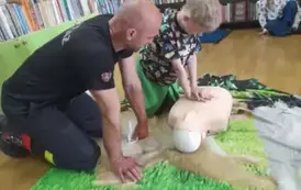 Chlopiec w blond włosach uciska klatkę piersiową fantoma, obok prezes straży się przygląda.