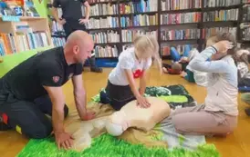 Dziewczynka w blond włosach uciska klatkę piersiową fantoma, obok siedzi druga dziewczynka i prezes straży.