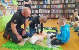 Na zielonym dywaniku chłopiec uciska klatkę piersiową fantoma, obok jest prezes straży i drugi chłopiec i się przyglądają.