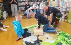 Na zielonym dywaniku chłopiec uciska klatkę piersiową fantoma, obok jest prezes straży i drugi chłopiec i sie przyglądają.