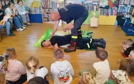 Dzieci siedzą na dywanie, a z przodu na dywaniku leży na boku pomocnik prezesa straży, prezes łapie go za ramię.