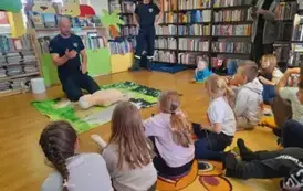 Dzieci siedzą na dywanie, z przodu na dywaniku siedzi prezes, a przy nim leży fantom. Po prawej stronie z przodu stoi jego pomocnik.