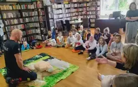 Dzieci siedzą na dywanie i siedziskach, na środku na dywaniku siedzi prezes OSP, a przy nim leży fantom.