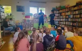 Dzieci siedzą na dywanie, z przodu po lewej stronie na trzech krzesłkach siedzi kobieta i dwie dziewczyny, z przodu stoi prezes OSP i jego pomoccnik.