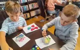 Dzieci siedzą przy stołach i malują farbami świeczniki z gipsu w kształcie serca.