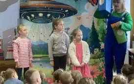 Przed dziećmi stoją na środku trzy dziewczynki i aktorka trzymająca kolorowe kosze do segregacji śmieci.