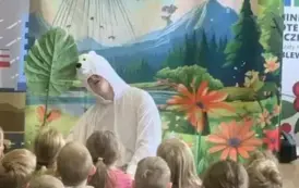 Przed publicznością dziecięcą siedzi aktor w przebraniu białego misia. W tle jest statek kosmiczny lądujący na ziemi i fruwające motyle.