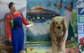 Przed dziećmi stoi pochylony aktor w przebraniu za psa, a obok aktor z lornetką i notesem w niebieskim kombinezonie. Wiszą u spodni mu zielone okulary.