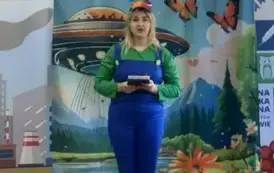 Przed dziećmi stoi aktorka w zielonej bluzce i w niebieskim kombinezonie trzymając w rękach notes. W tle statek kosmiczny na ziemi i fruwające motyle.