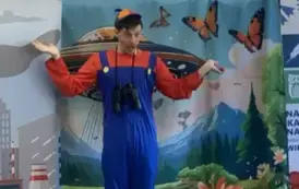 Na tle statku  kosmicznego na ziemi stoi aktor w czerwonej bluzce i niebieskim kombinezonie, ma rozłożone ręce w bok, a na szyi wisi lornetka.