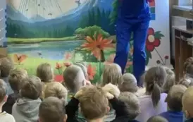 Dzieci patrzą do przodu, a tam aktor ubrany w czerwoną bluzkę i niebieski kombinezon patrzy przez lornetkę. W tle statek kosmiczny na ziemi i fruwające motyle.